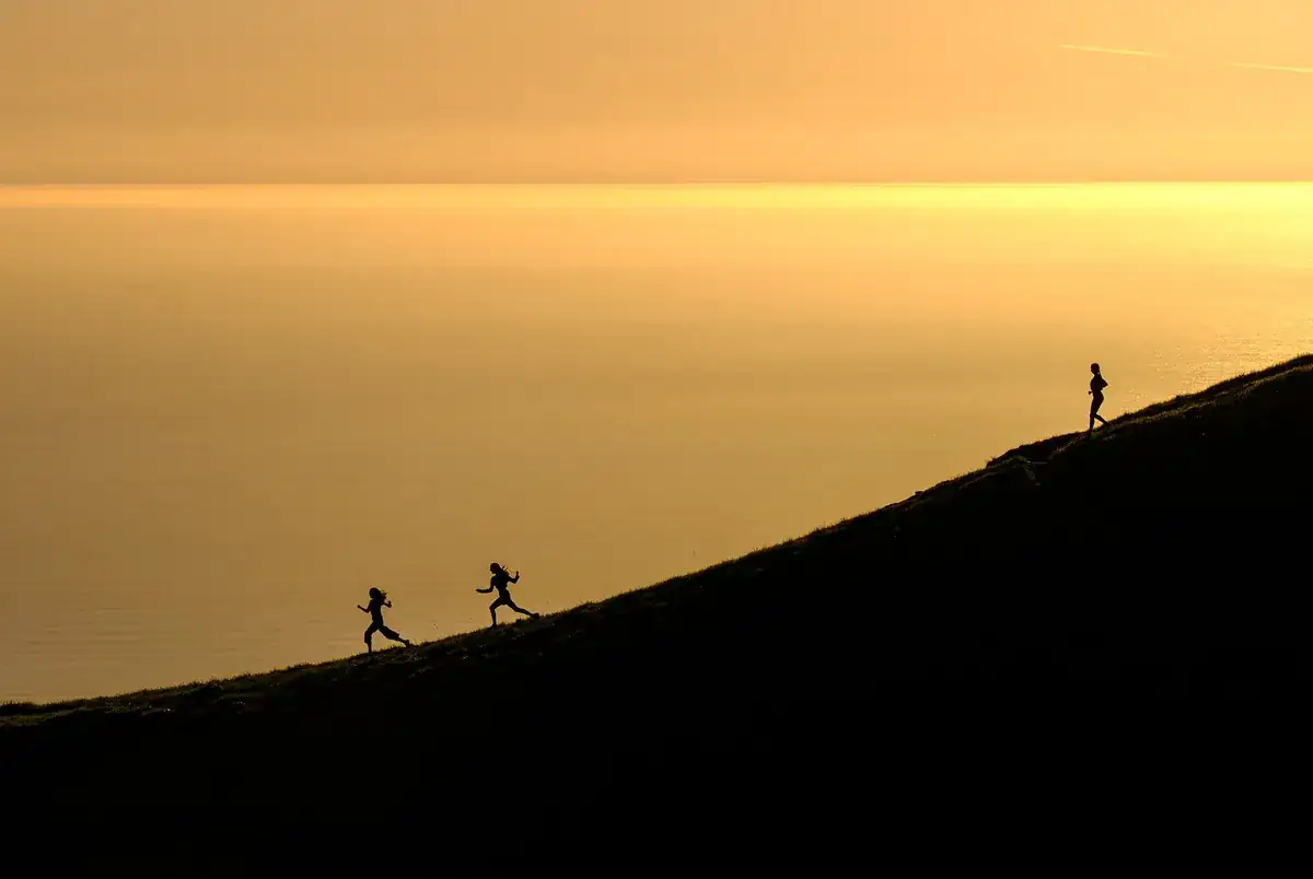 Silhuetas de duas pessoas a correr numa encosta contra um céu ao amanhecer.